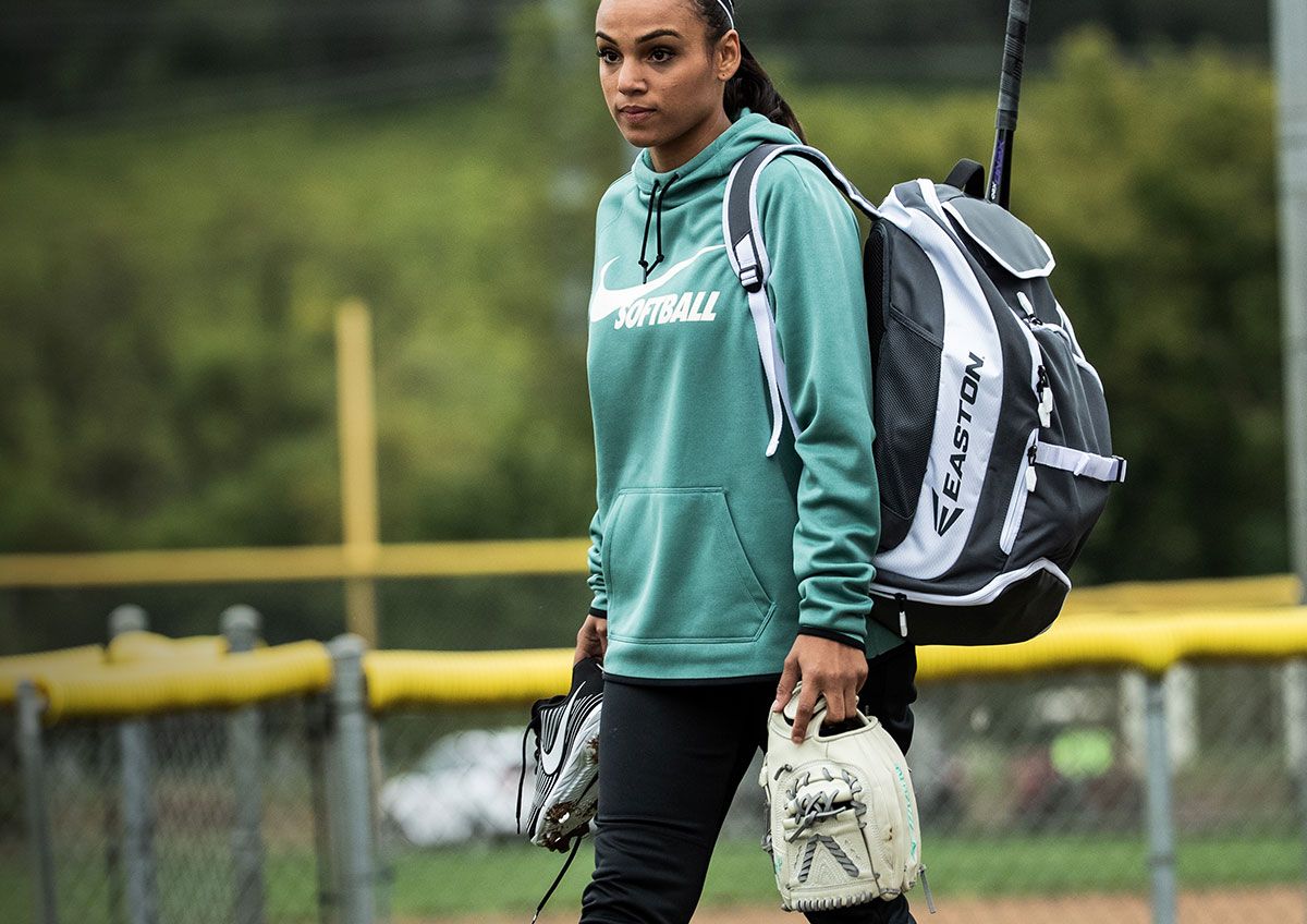 A young woman on a baseball field carrying her baseball gear, wearing a Nike Women's Therma Pullover Softball Hoodie.