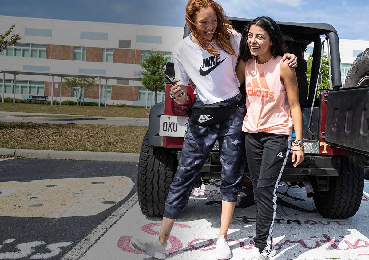 Girl In Nike Shirt And Nike Pants Hugging Girl In Adidas Shirt And Adidas Pants